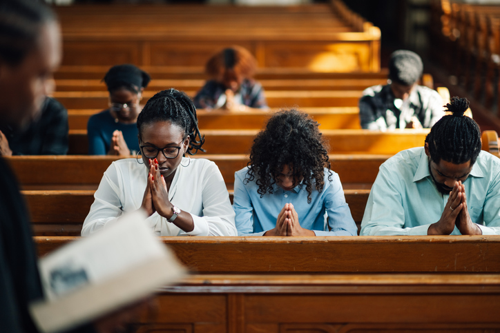 Black church members sitting in pews with heads bowed in prayer