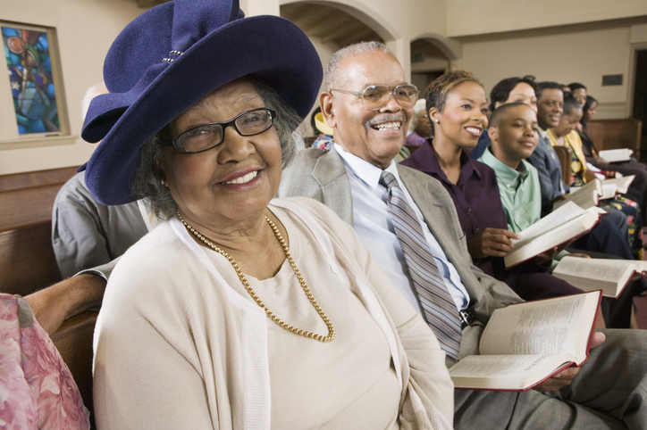 Elderly Black woman smiling in church pew surrounded by family and friends holding hymnals